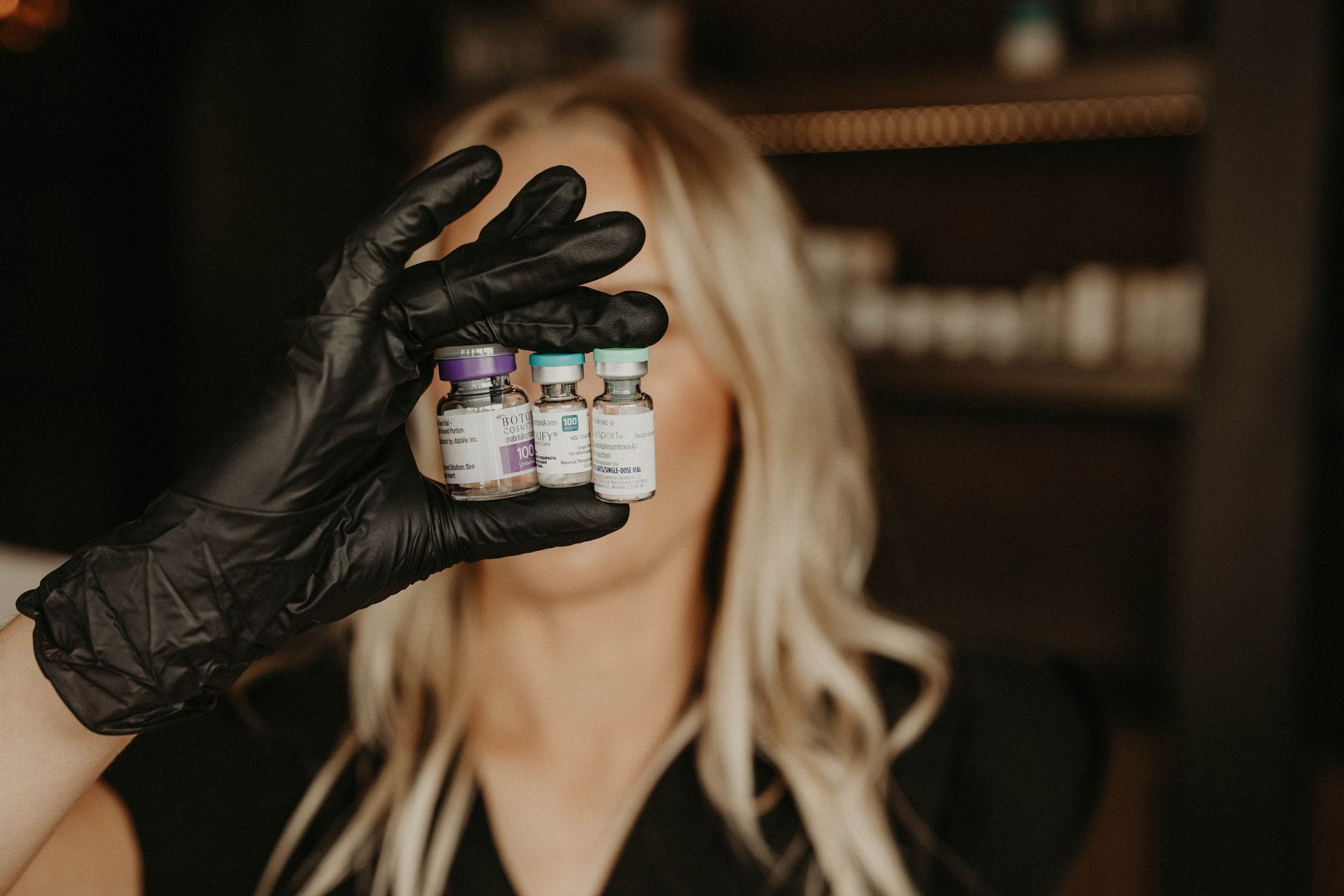 Black-gloved hand holding three small vials in front of a blurred blonde woman in a dark interior
