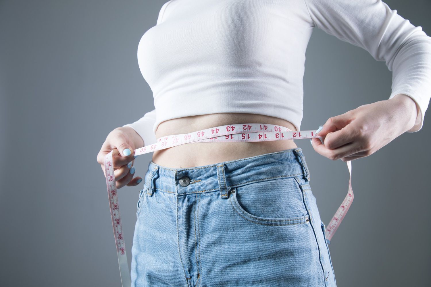 Woman in white top and jeans measuring waist with a tape measure.