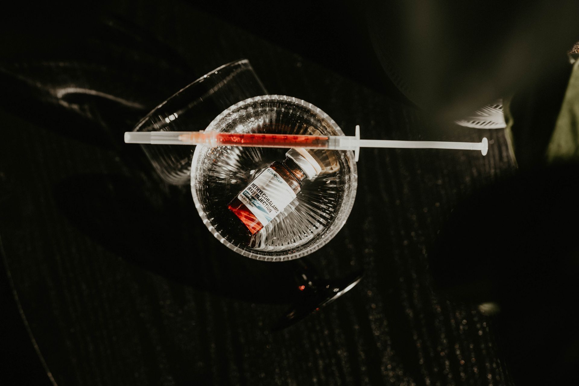 Syringe resting on a glass dish containing a vial with red liquid; black background.