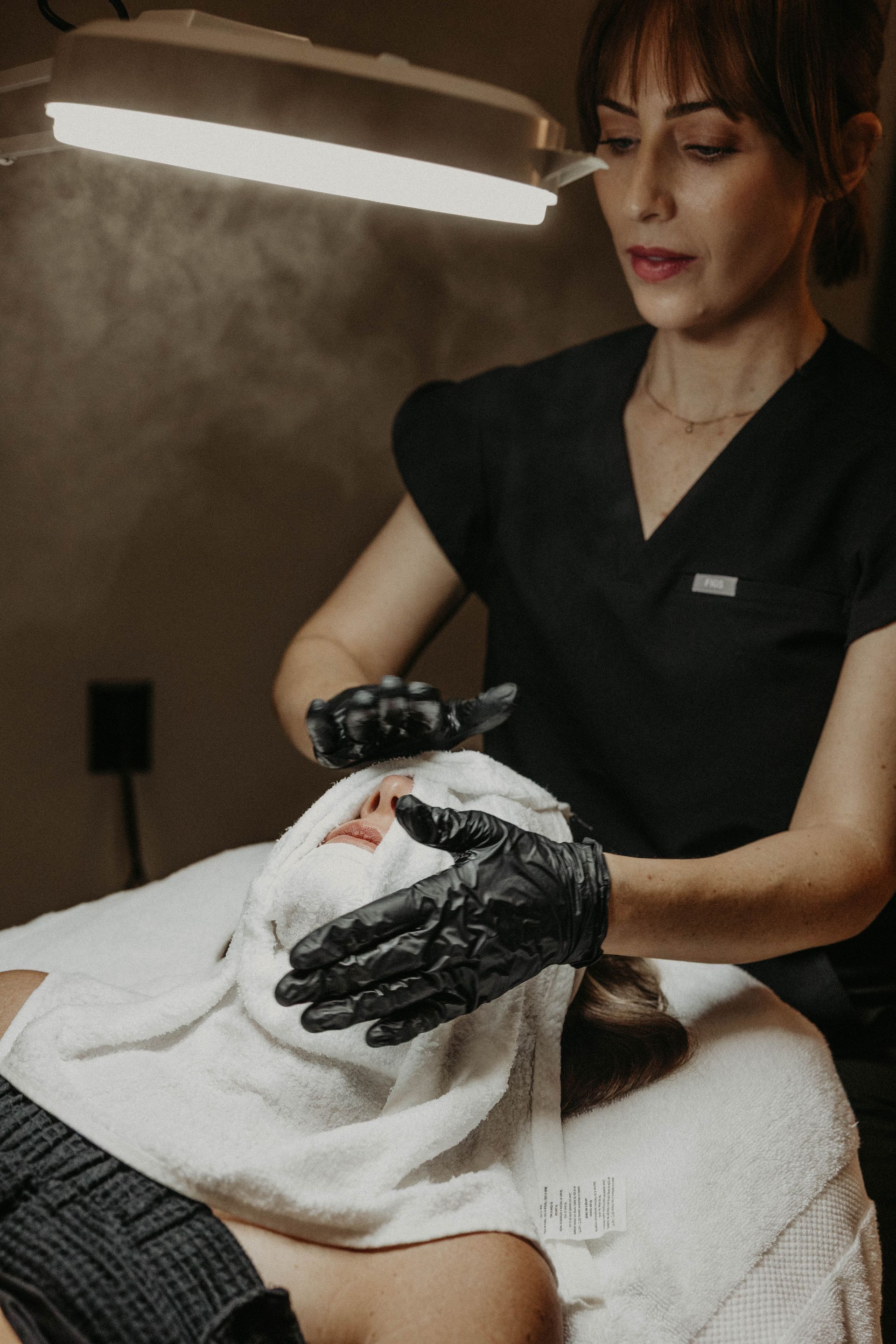 Woman giving facial treatment: gloved hands on client's face covered with towel. Spa setting, warm light.