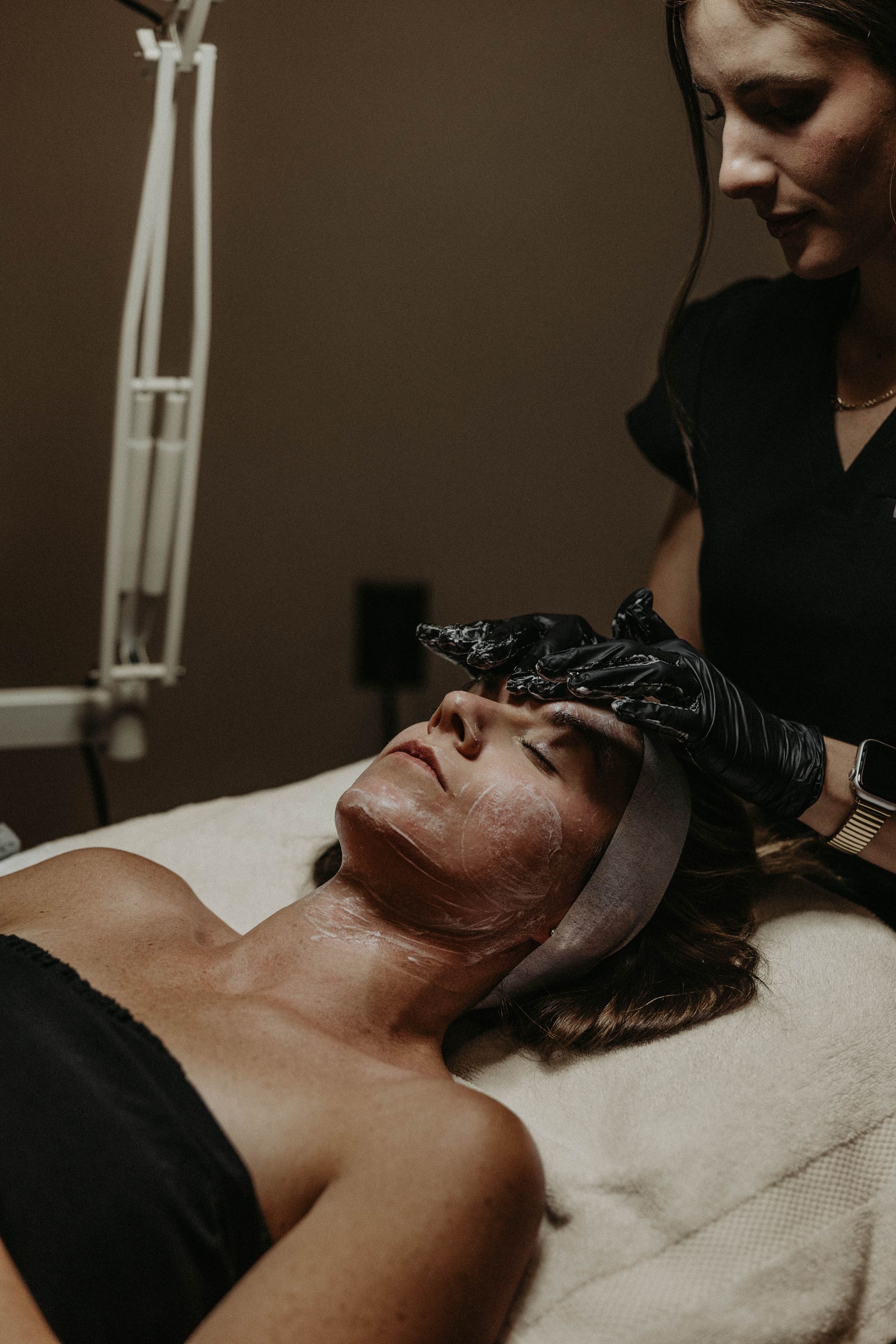 Woman receiving facial treatment from esthetician in a salon; both are wearing black clothing.