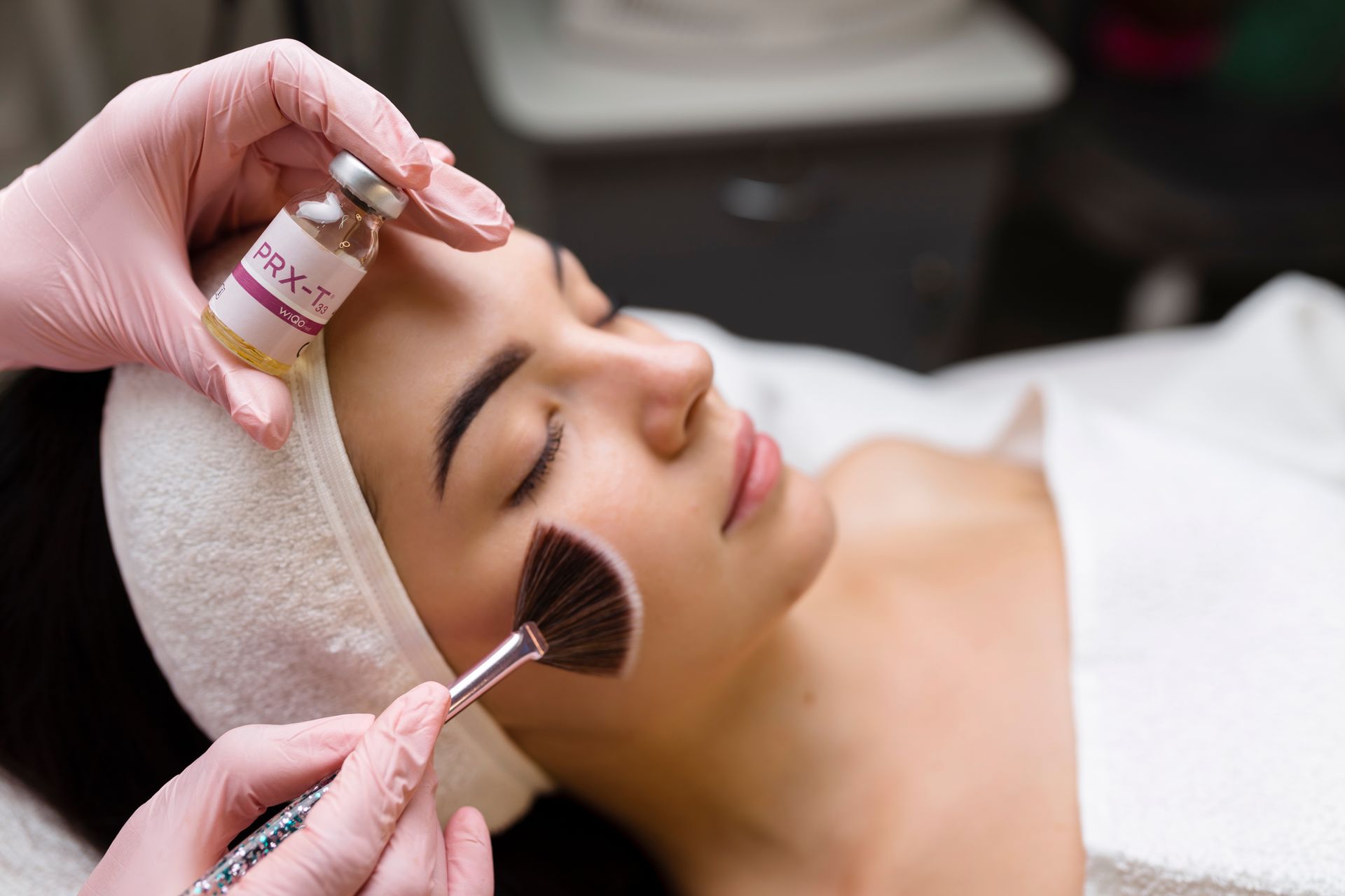 A woman receives a facial treatment. A gloved hand holds a bottle, the other applies product with a brush.