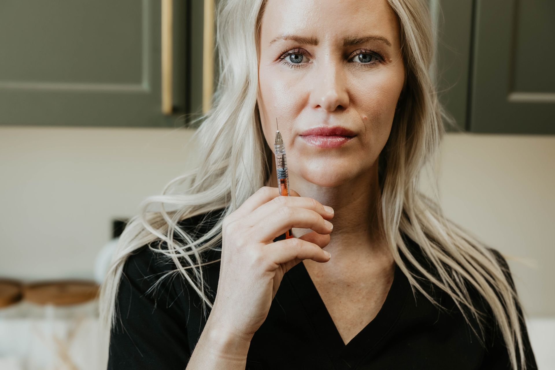Woman holding syringe, looking at camera, potentially preparing for an injection. Light hair, black top.