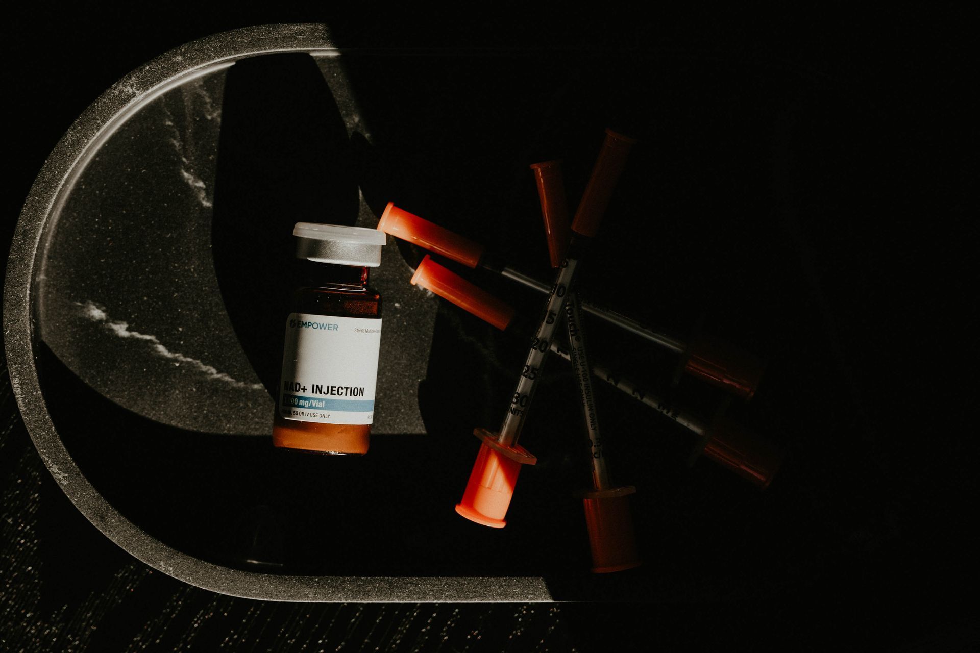 Vial of medication with needles in a dark setting, orange and white labels visible, in a tray.