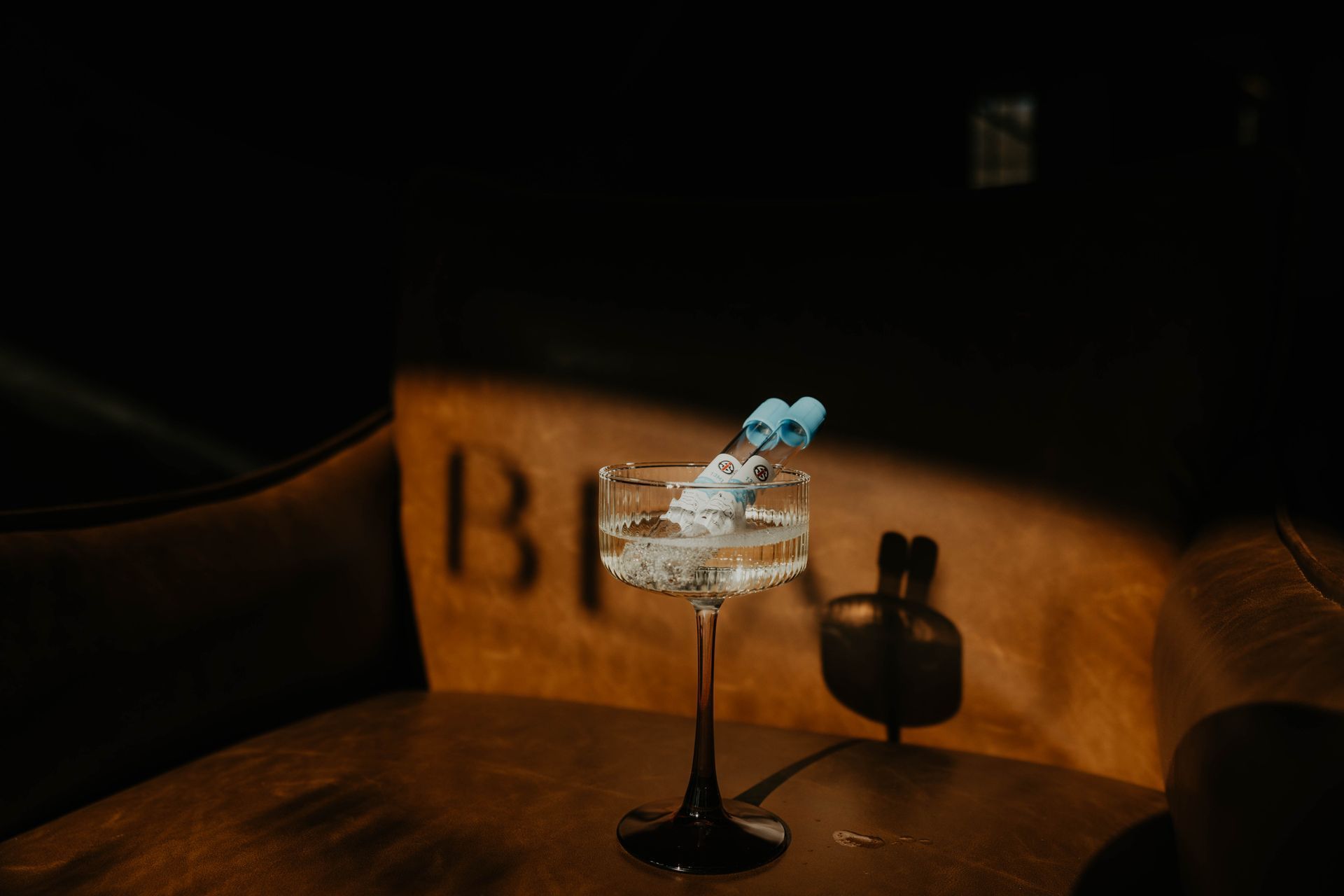 Cocktail in stemmed glass, garnished with blue decorative elements, on a leather chair. Dark, moody setting.