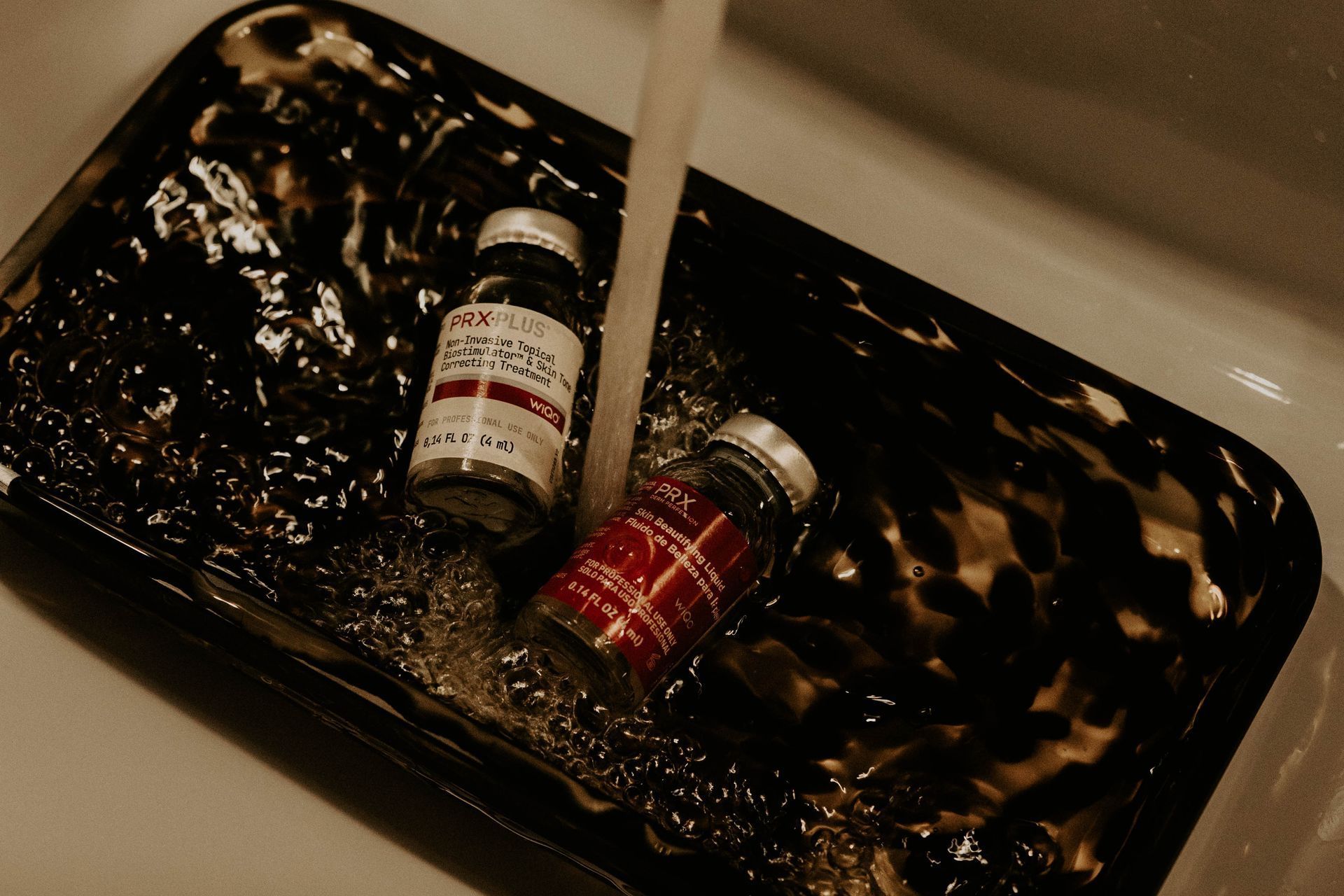 Two medicine vials in a tray being rinsed with water in a sink.