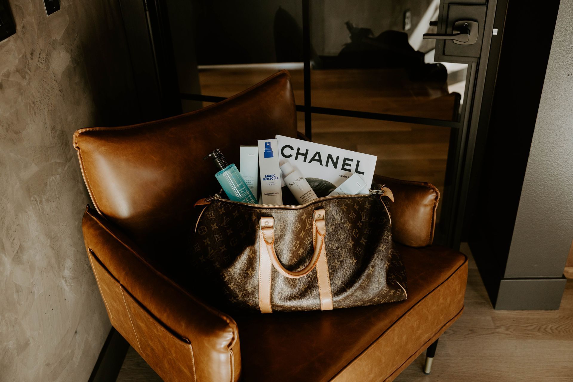 Brown leather chair with Louis Vuitton bag filled with beauty products and a Chanel magazine.