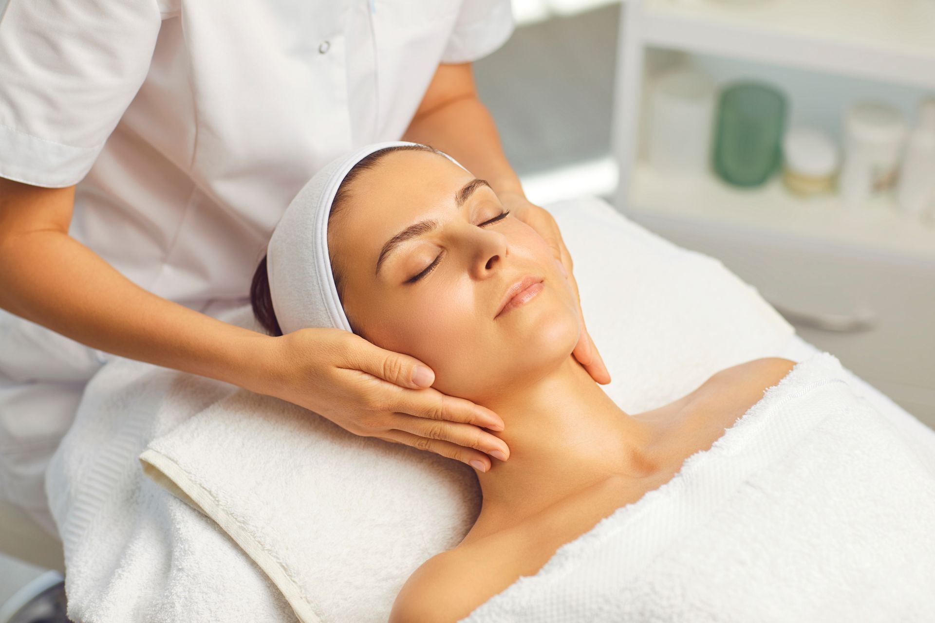 A spa facial treatment with a woman lying on a towel while a therapist massages her face and neck
