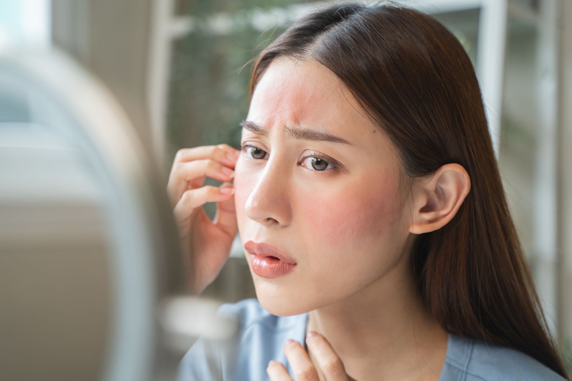 Person applying makeup near a mirror, touching an eyebrow with a concerned expression