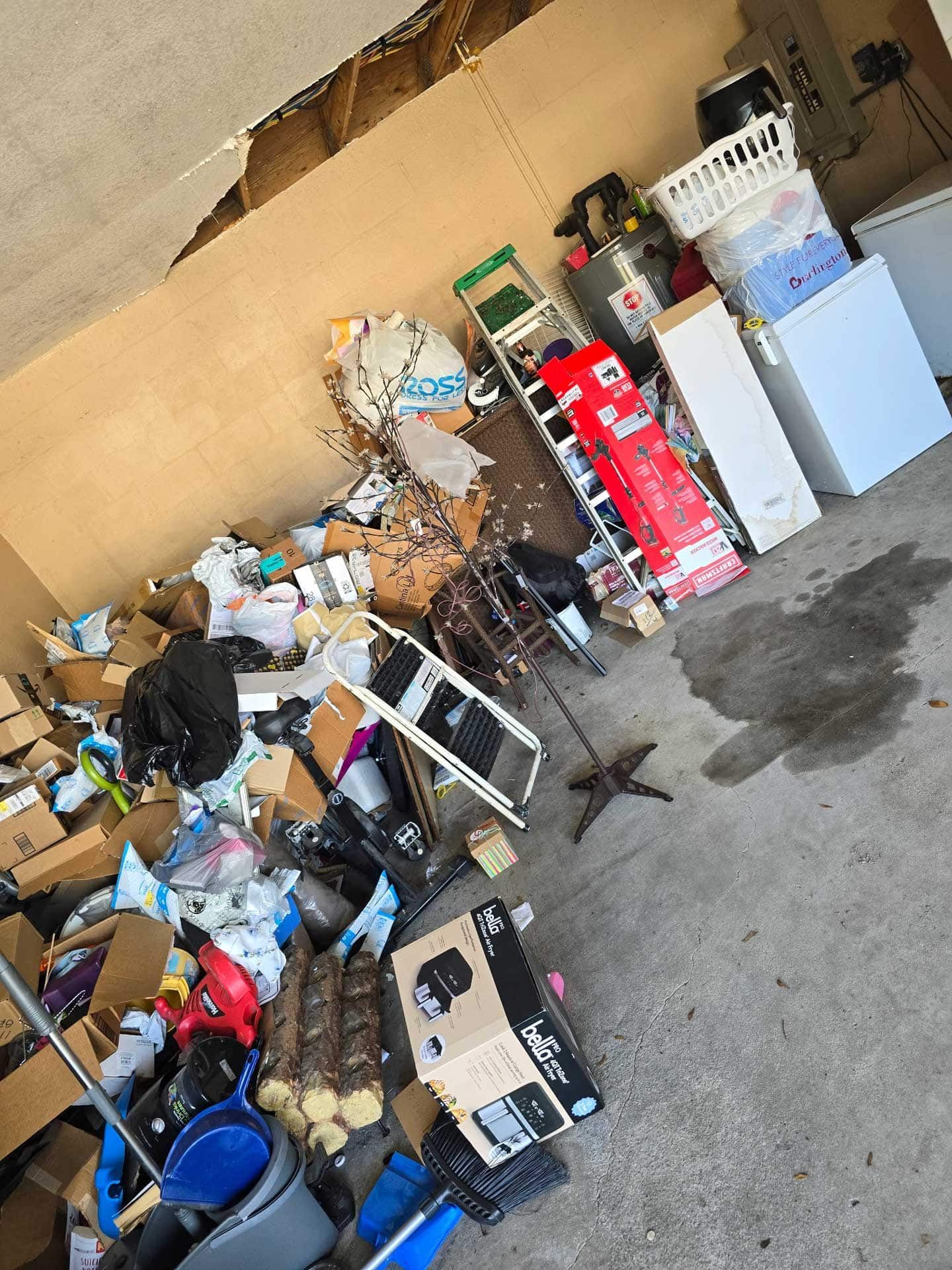 Cluttered garage with boxes, trash, ladder, and various items. Ceiling damage and water stains are visible.