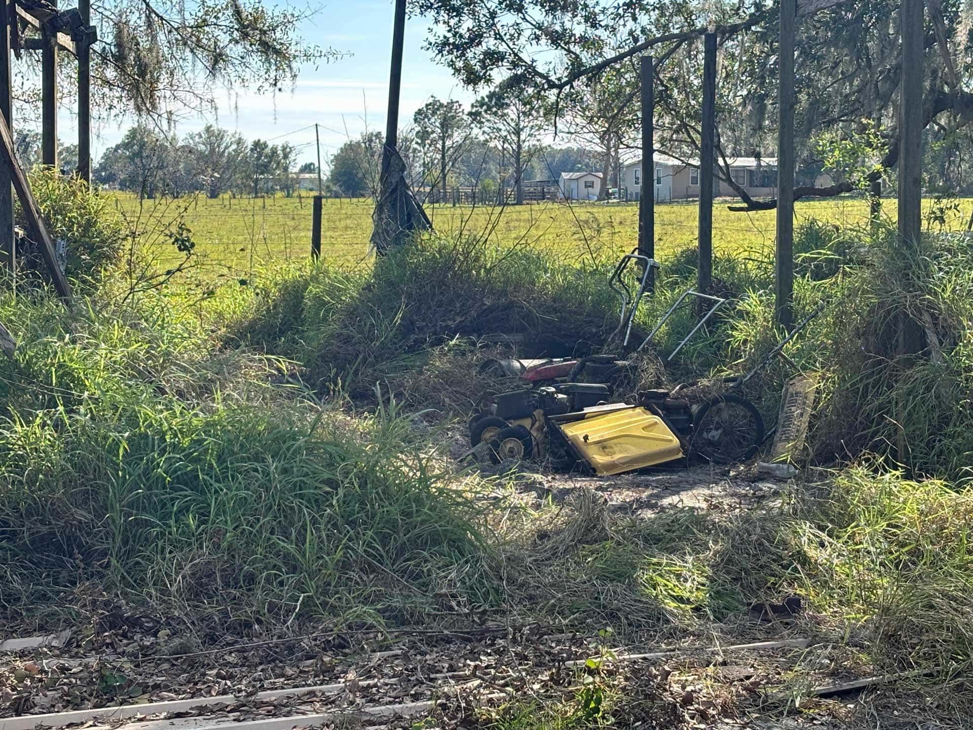 A small, yellow lawn mower sits in tall grass near a fence, surrounded by a field and trees.