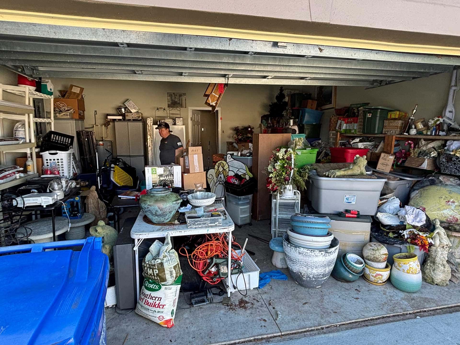 Garage sale with cluttered items: tables, bins, pots, and two people.