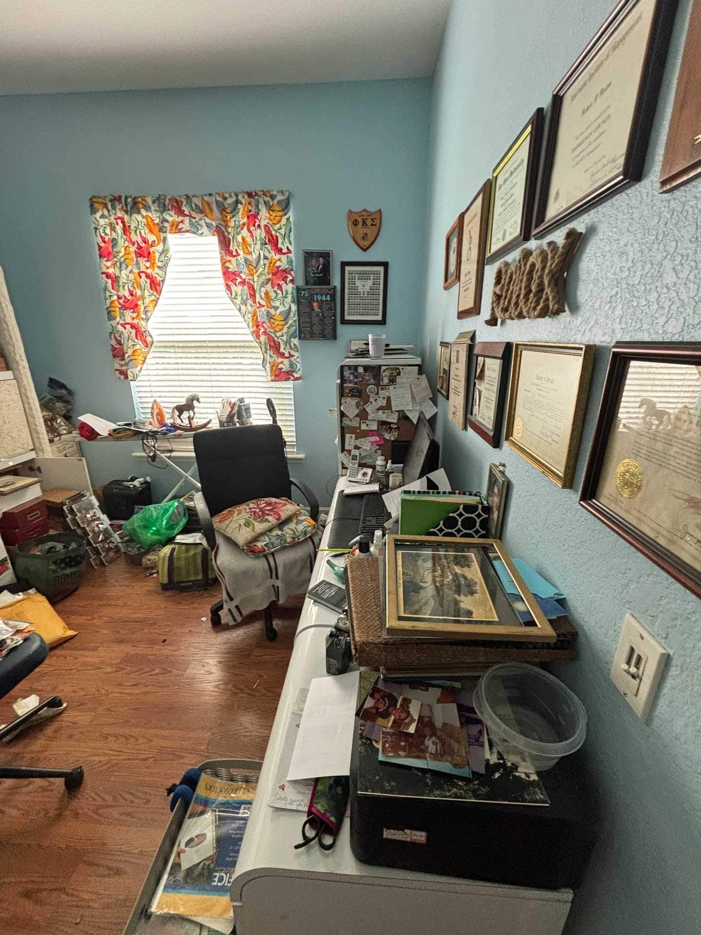 Cluttered office with a desk, chair, window, and wall-mounted certificates. Light blue walls, floral curtains.