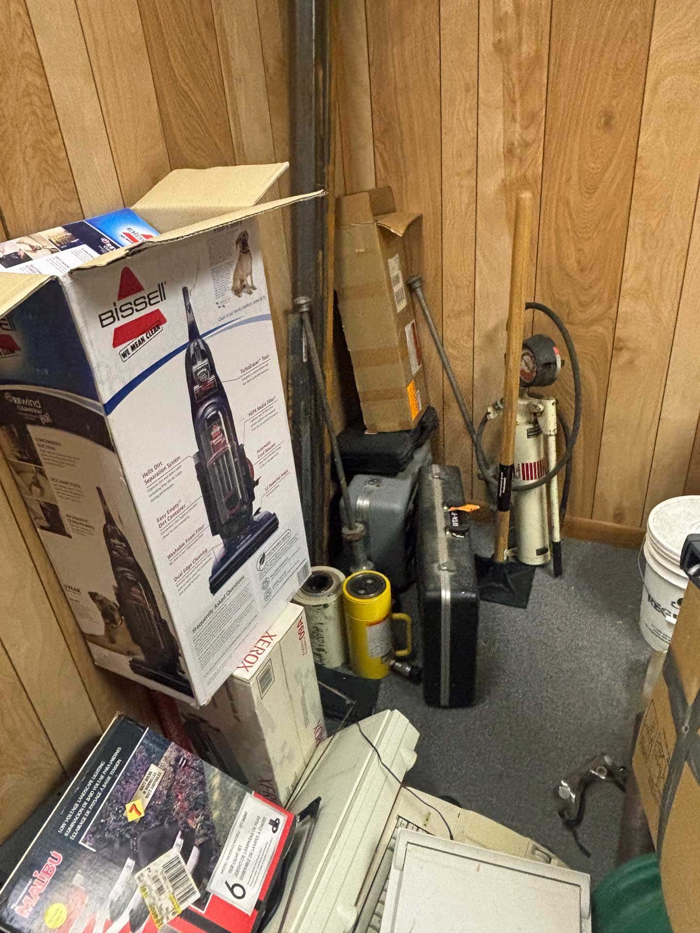A cluttered storage area with a Bissell carpet cleaner box, tools, and other items against wooden paneling.