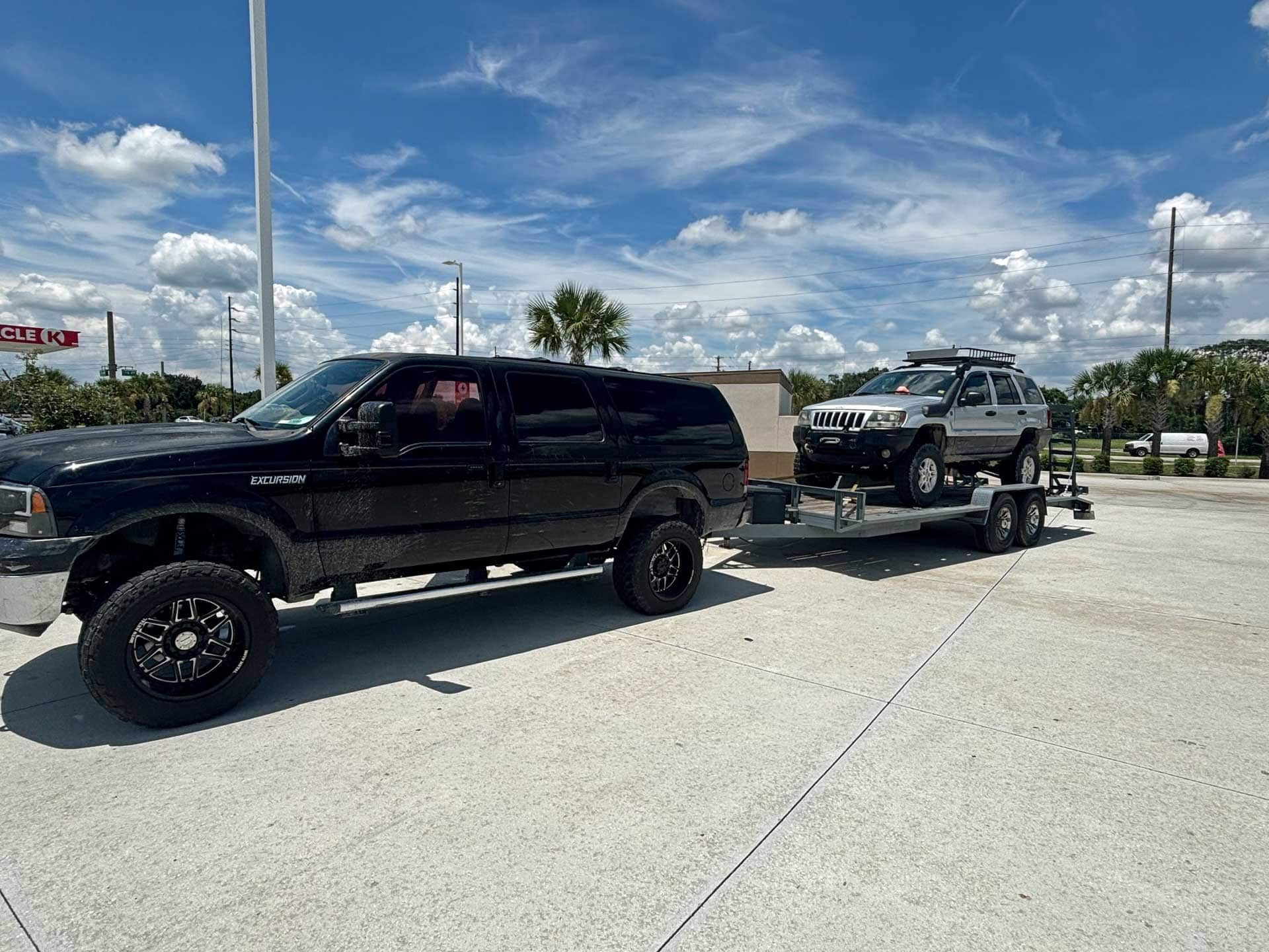 Black SUV towing a silver SUV on a trailer under a blue sky.