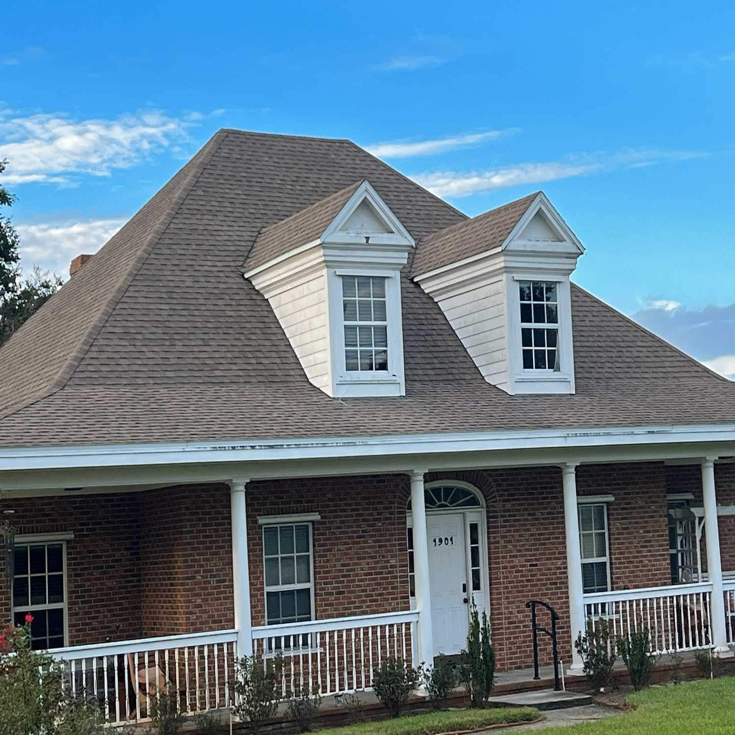 Brown brick house with white trim, porch, and two dormers under a brown shingle roof, set against a blue sky.
