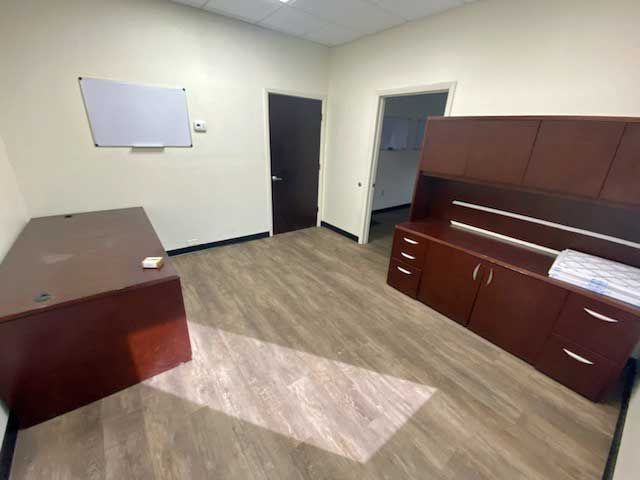 Empty office with desk, cabinet, whiteboard, and open doorway, wood-look floor.