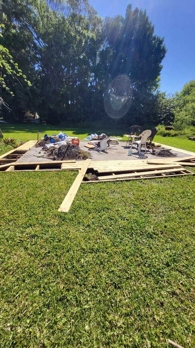 Construction site in grassy yard; wooden frame with debris, tools, and materials. Bright sunlight, trees in the background.