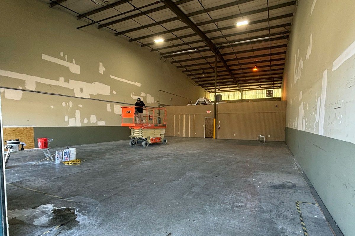 Interior of a warehouse under construction, with a worker on a lift. Concrete floor, green and tan walls, metal ceiling.