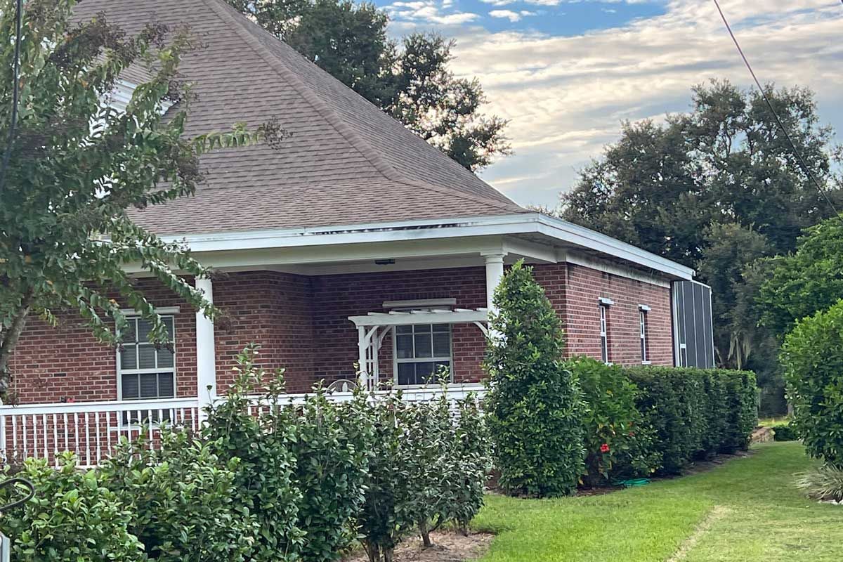 Red brick house with a porch and bushes, surrounded by trees. Cloudy sky.