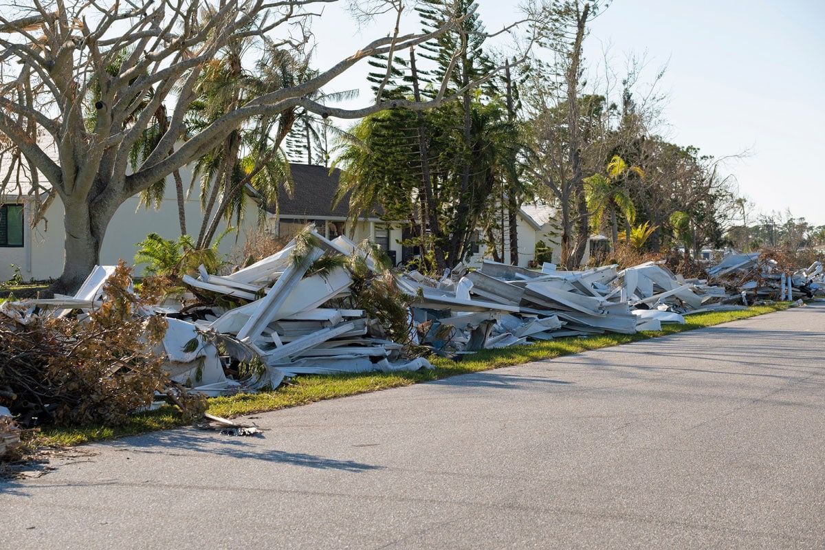 Debris piled along a road next to a damaged house and trees after a storm.