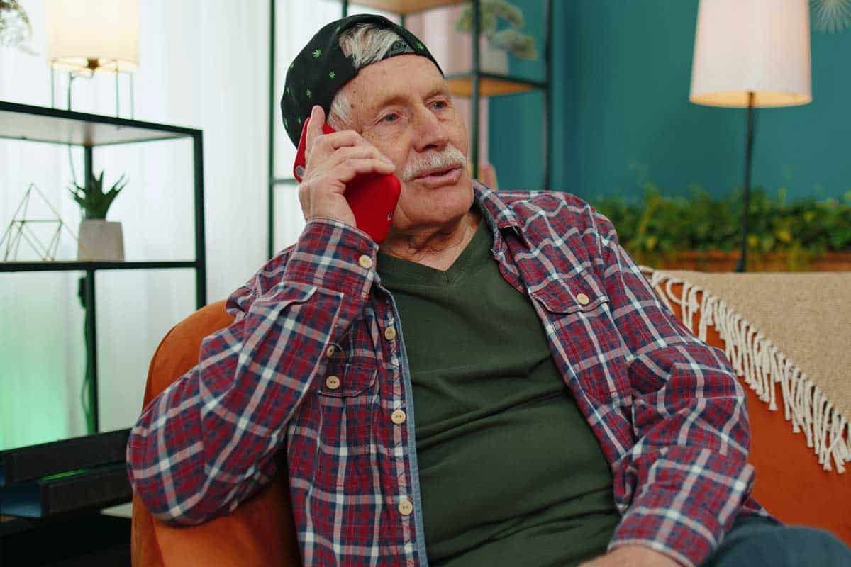 An older person in a plaid shirt talks on a red phone, sitting on an orange couch.