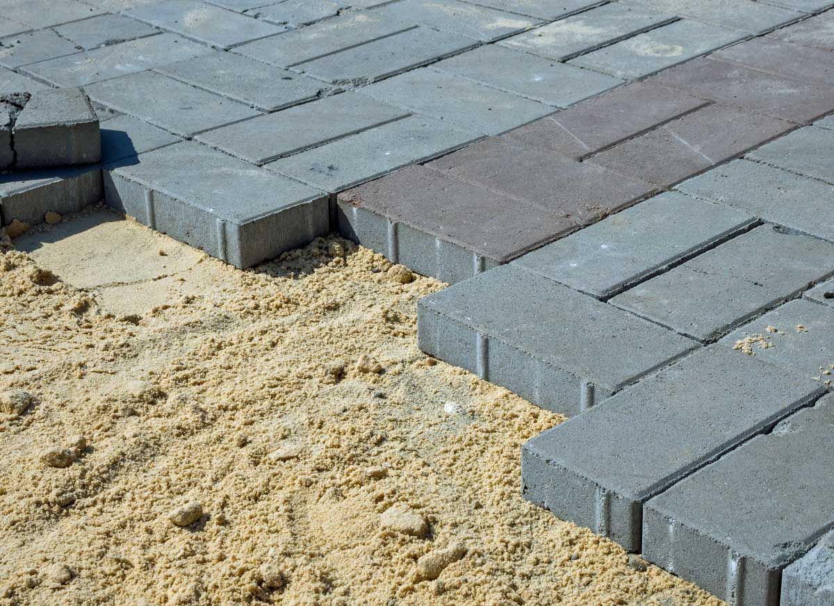 Gray brick pavers being laid on a bed of sand; some pavers installed, others in progress.