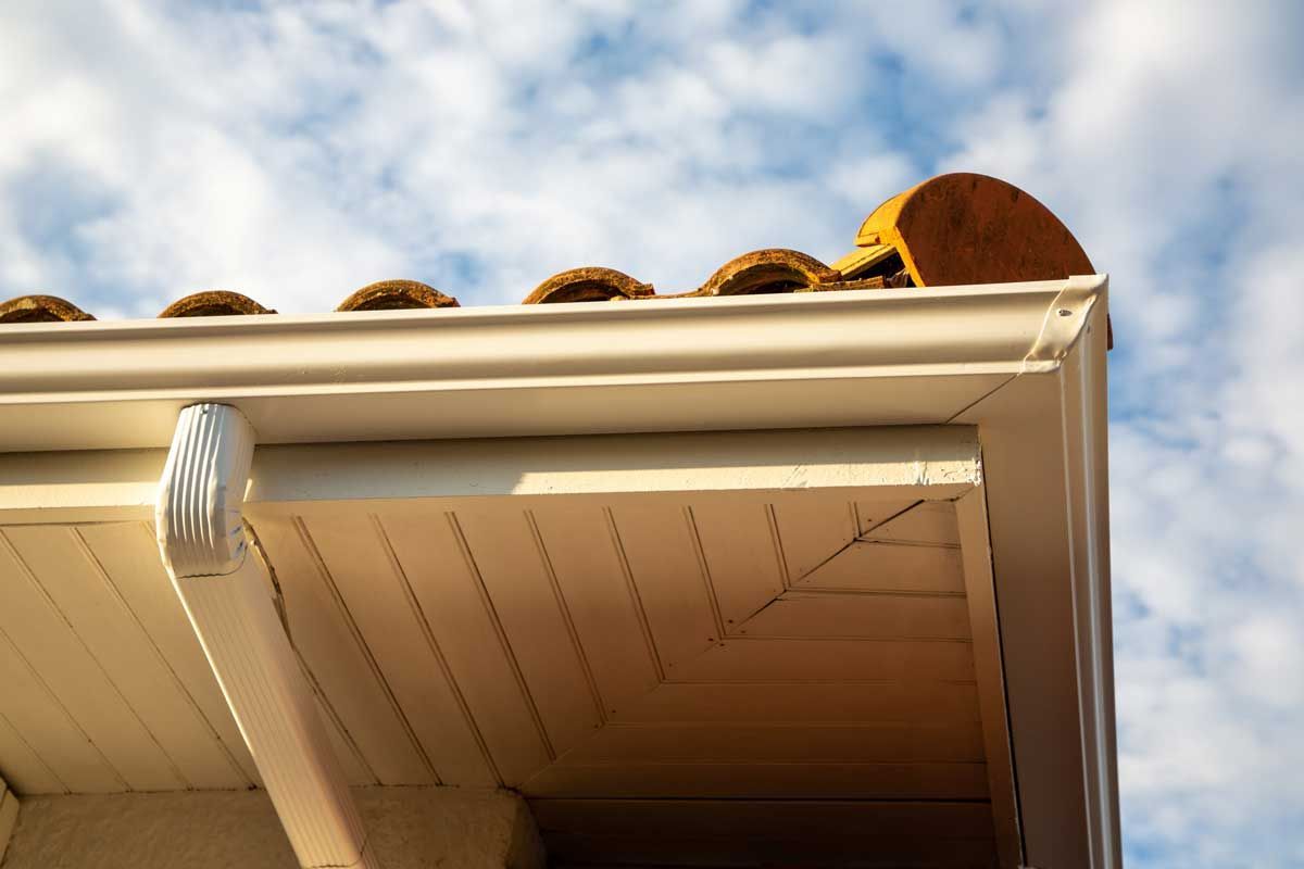 Beige rain gutter on a house, with terracotta roof tiles and a blue sky in the background.