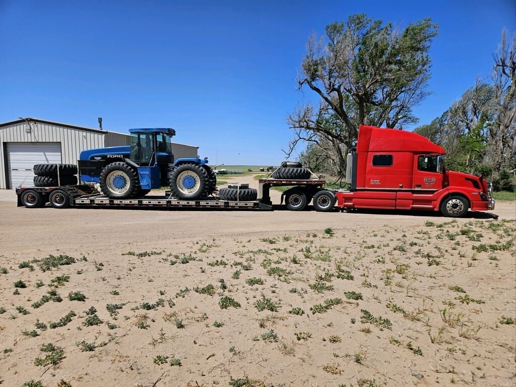A red semi truck is carrying a blue tractor on a trailer.
