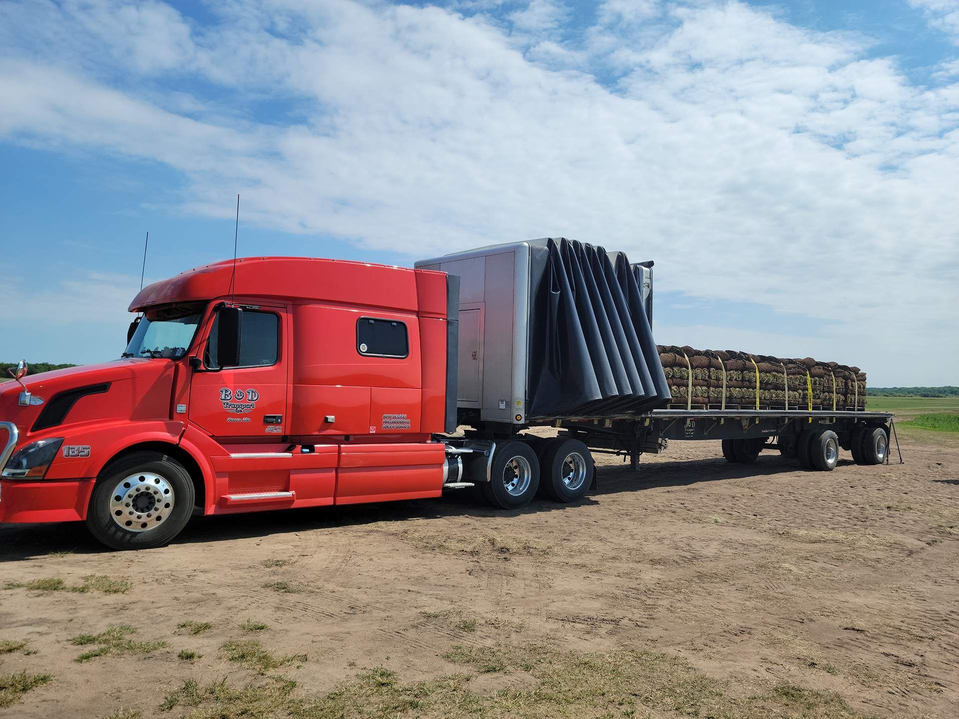 A red semi truck is carrying a blue tractor on a trailer.