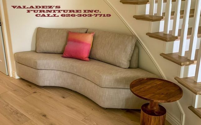 Curved beige bench under stairs with pink/orange pillow and small wooden table.