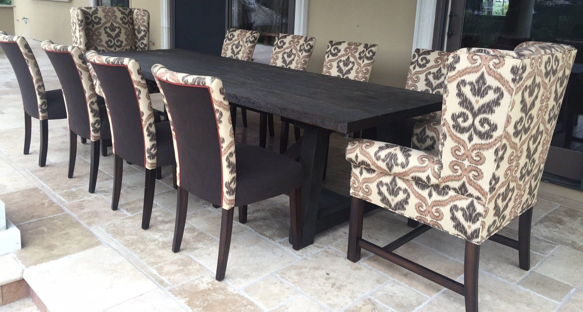 A long wooden dining table with 10 upholstered chairs on a stone patio.