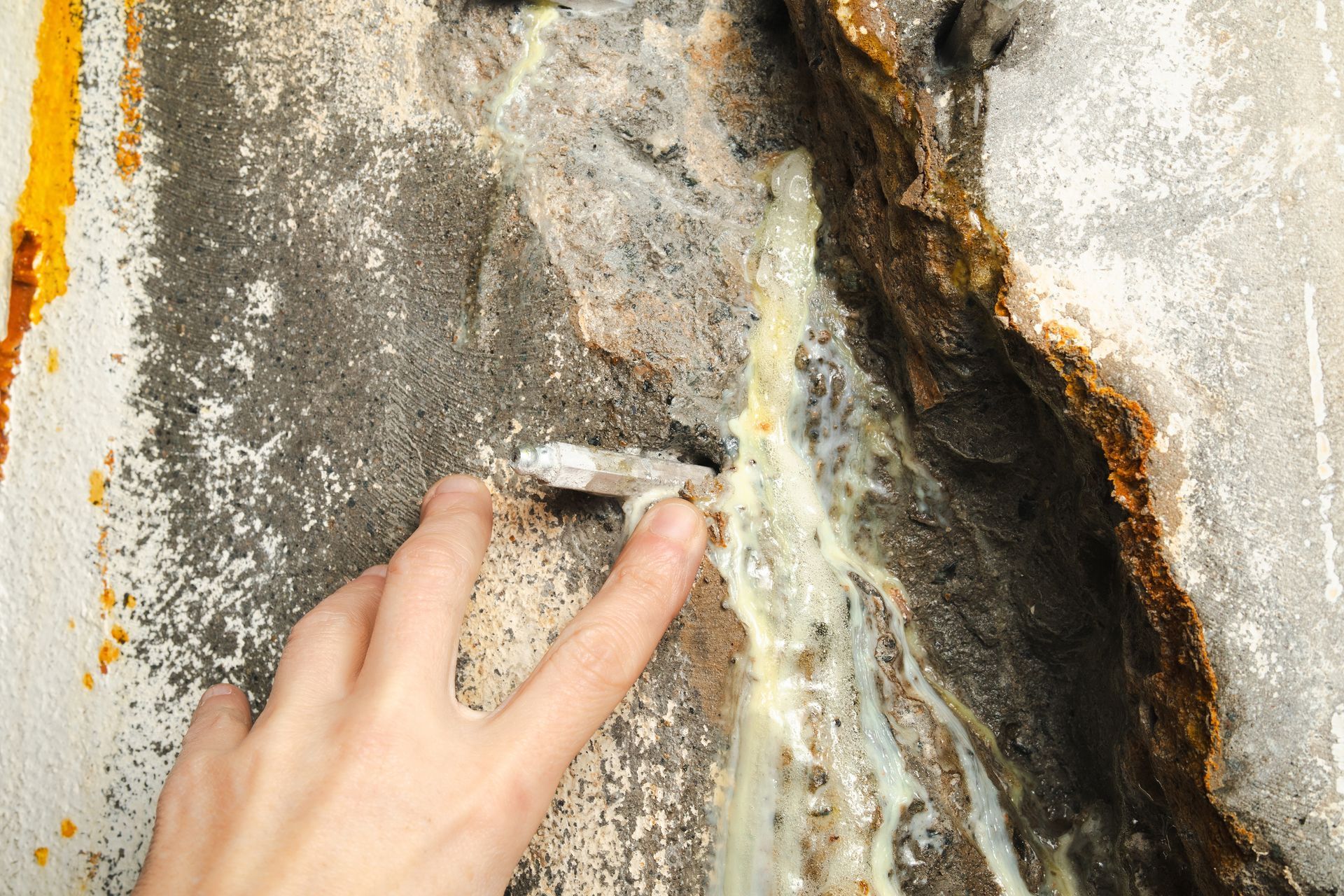 Hand touching a cracked, weathered concrete wall with peeling paint and exposed rust-stained damage