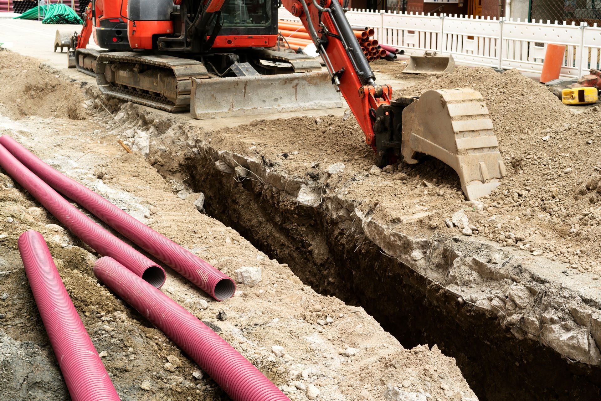Excavator digging a trench beside pink utility pipes at a construction site