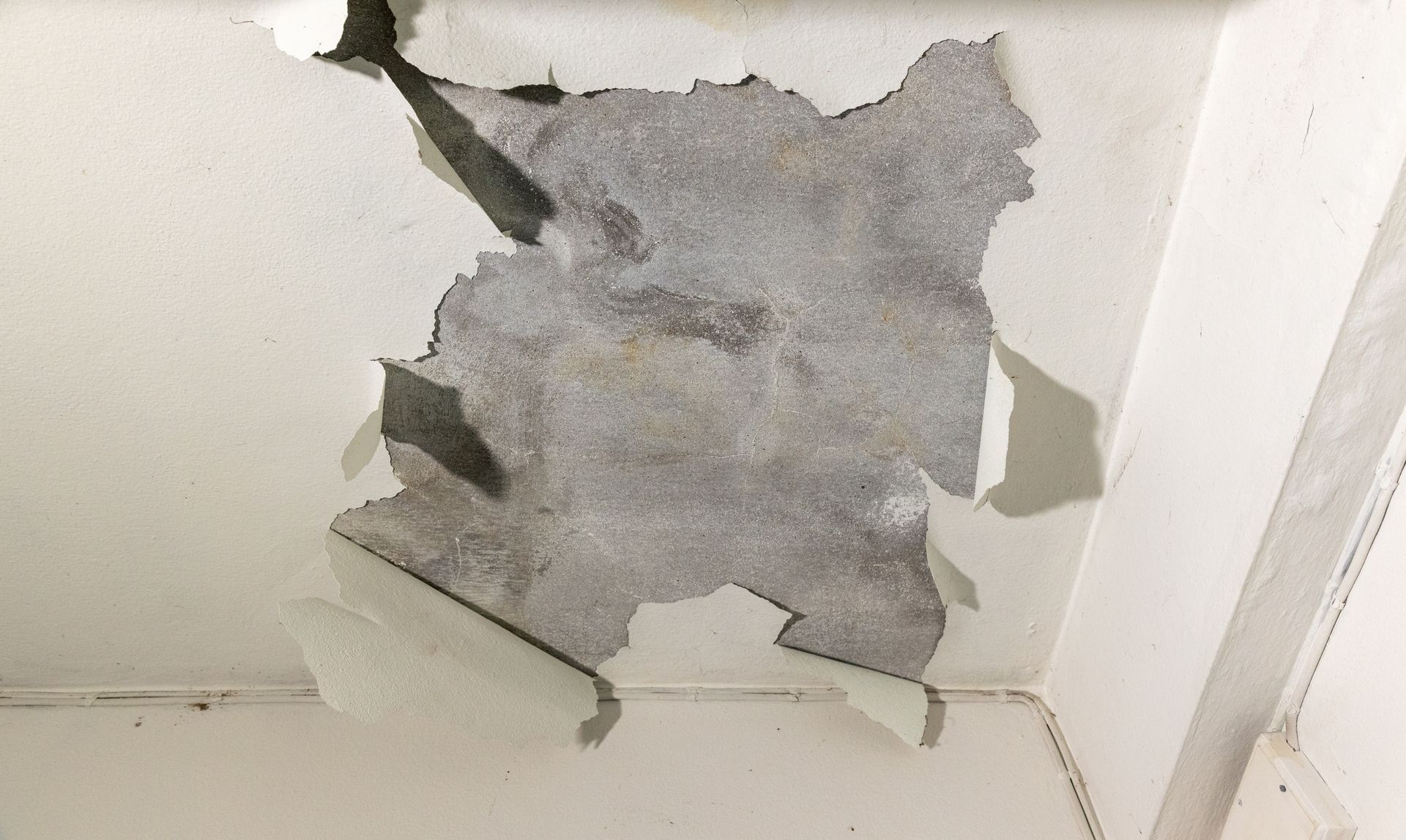 Damaged ceiling with a large gray patch of peeling plaster above a white wall corner