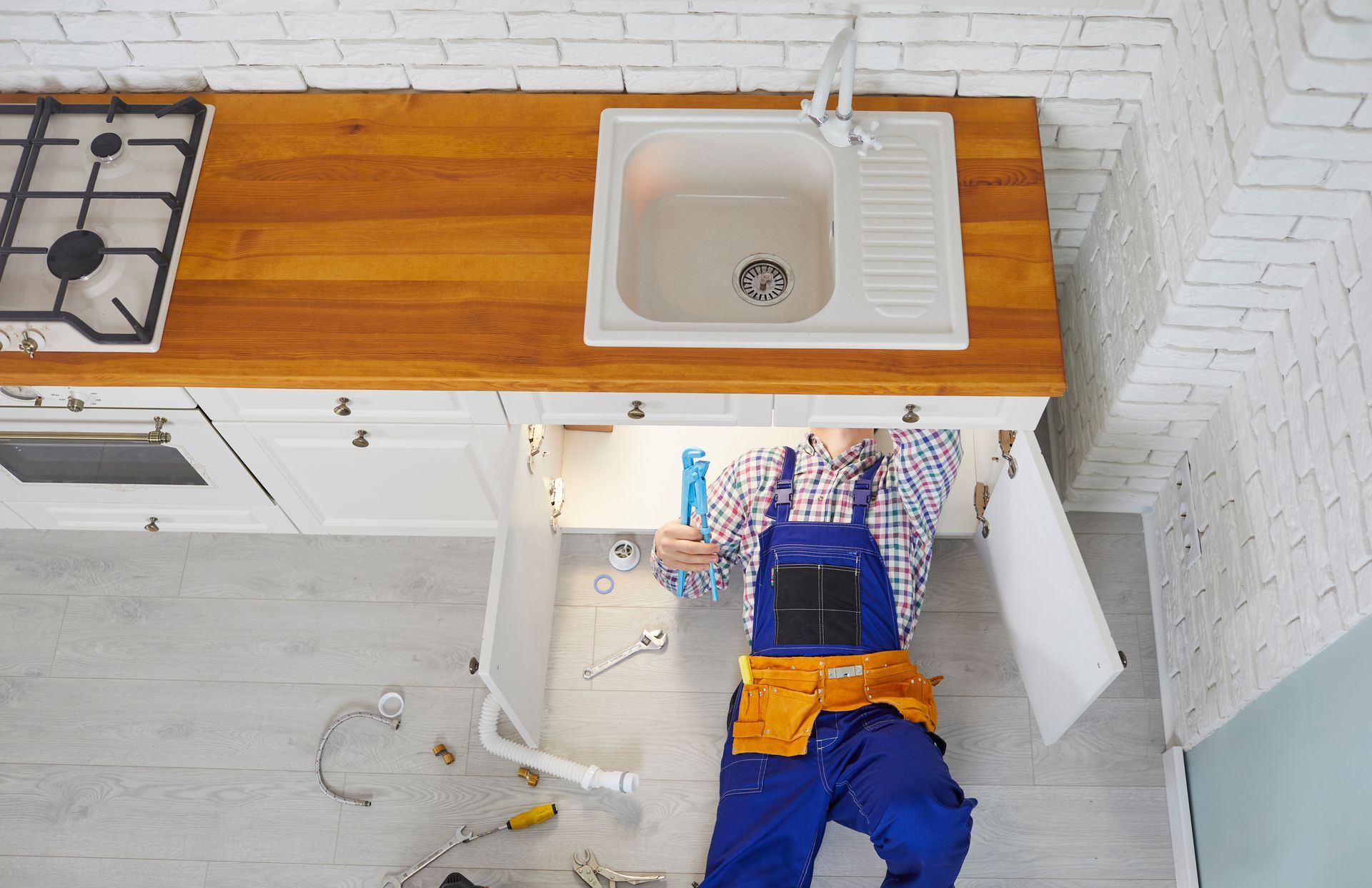 Plumber in blue overalls working under a kitchen sink, overhead view.