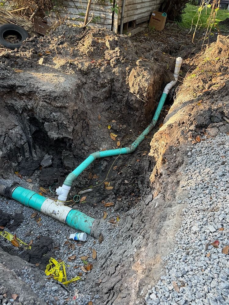 Trench with exposed green pipe and white fittings in a dug-out dirt yard near a fence.