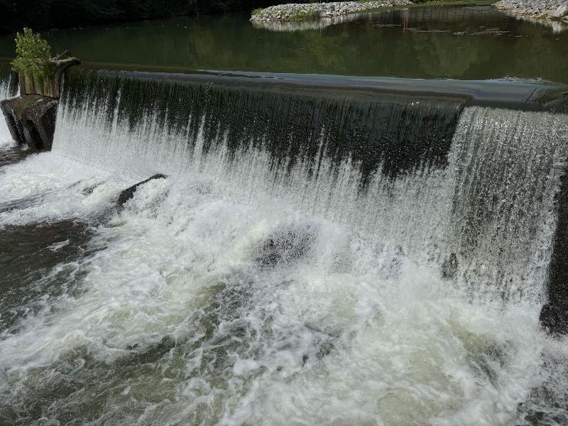 Water cascades over a dam, creating white foamy water below. A green treeline is visible in the background.