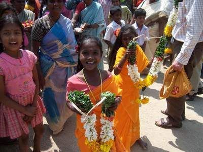 Children in orange dresses hold garlands, smiling, surrounded by people in India.