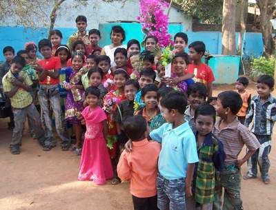 Group of children smiling, posing outdoors; some hold flowers, wear colorful clothes.