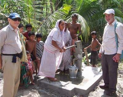 People drawing water from a well in a village. Woman pumps, others watch.