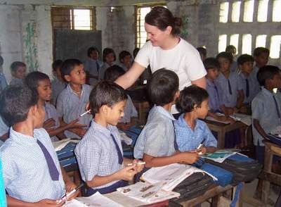 Teacher assists students in a classroom; children in uniforms, desks, and open books present.
