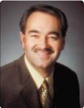 Man with dark hair, mustache, and brown suit smiling.