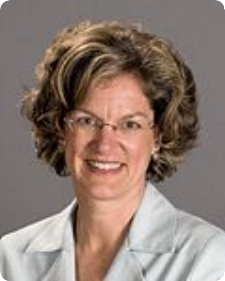 Woman with glasses, smiling, in a light blue jacket, against a gray background.