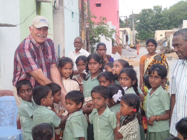 Man with children, presumably in India, smiles at the group. Children wear green uniforms, buildings are in the background.