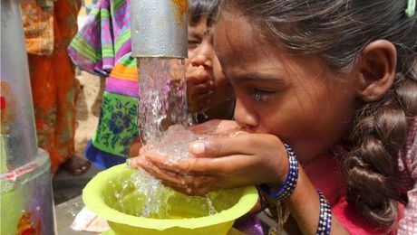 Girl drinking clean water from a tap with a yellow bowl, outdoors, other people visible.