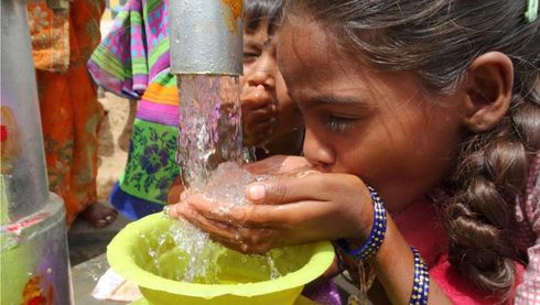 Girl drinking water from a tap, pouring into a bowl, others behind.