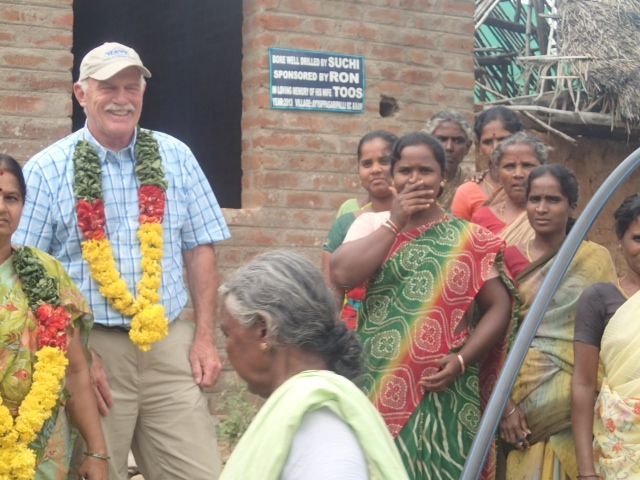 Man in a floral garland with women in saris. They stand near a brick building; several women smile.