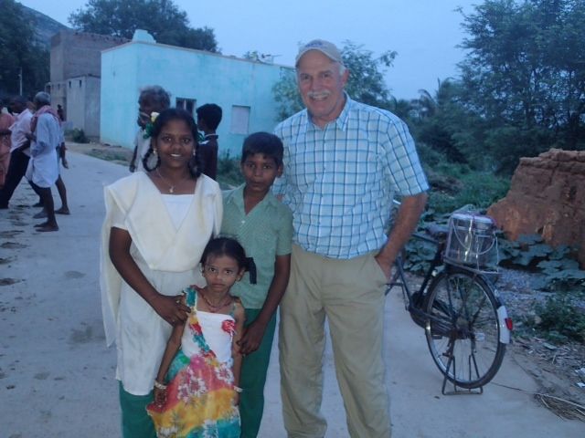 Man poses with three children in India, near a bicycle and buildings.