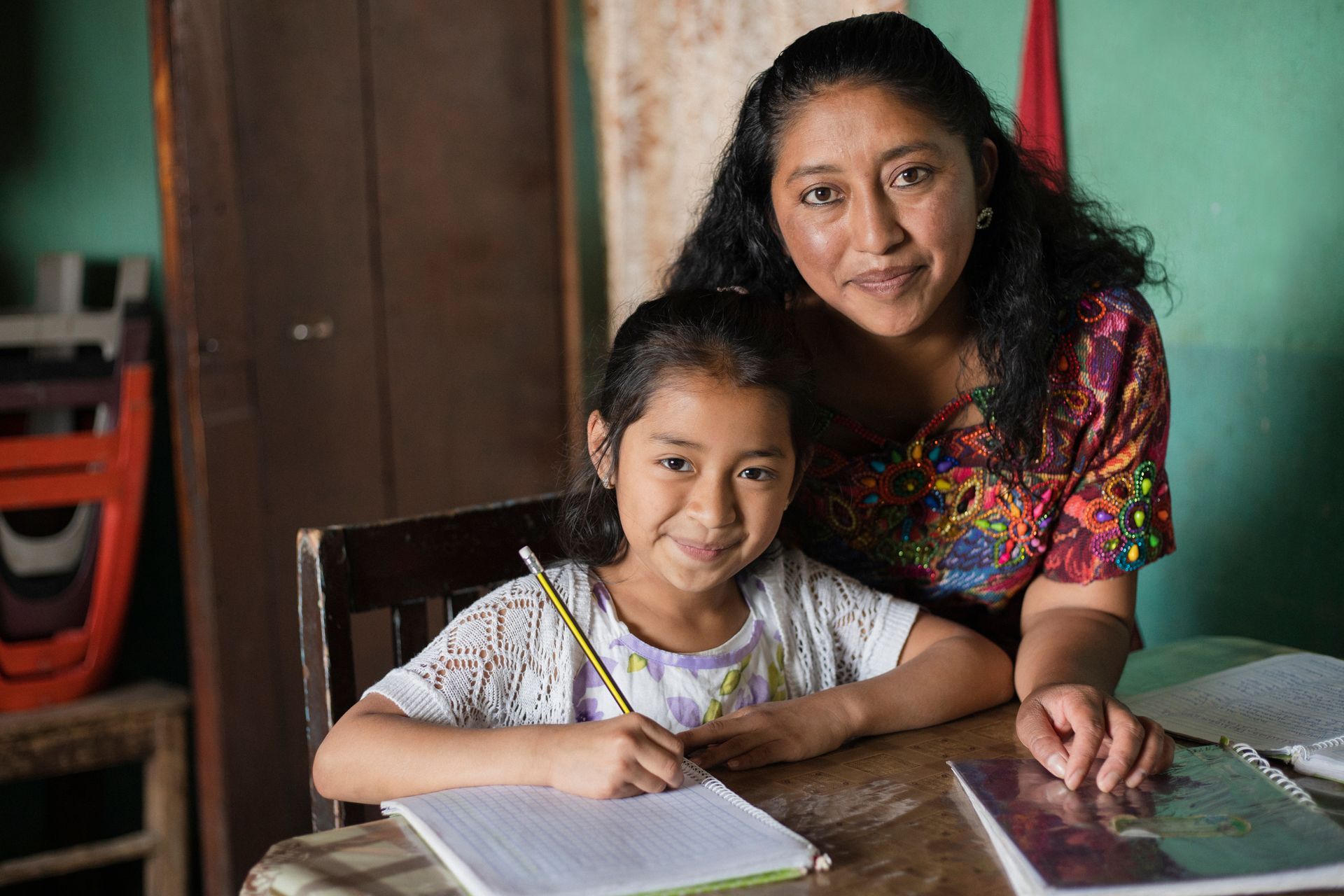 Woman helps a girl with homework, both smiling. They sit at a table in a home.